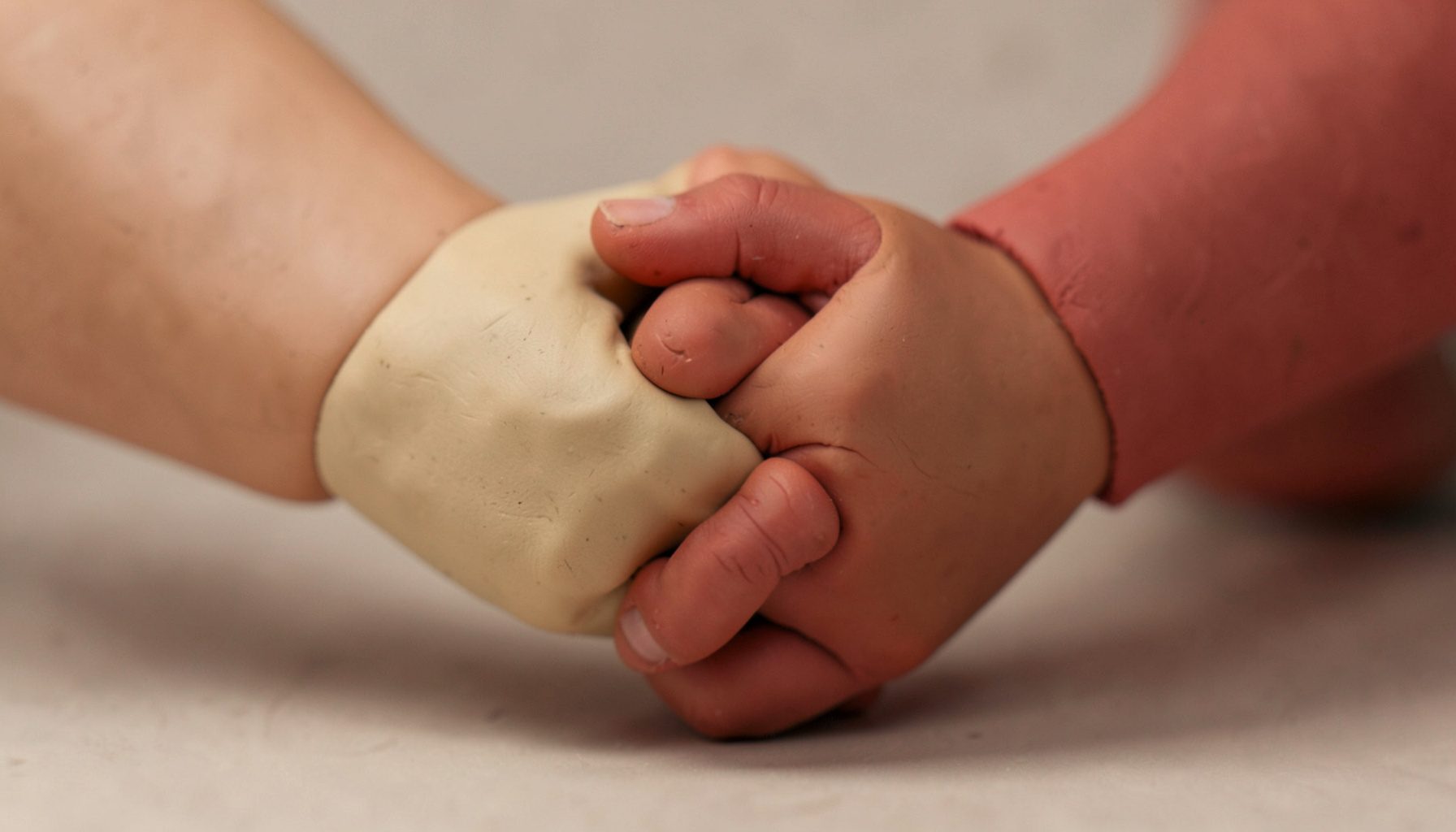 A softly lit clay-model scene of two hands clasped together in support, warm beige and muted terracotta tones, rounded handmade forms conveying unity and resilience.